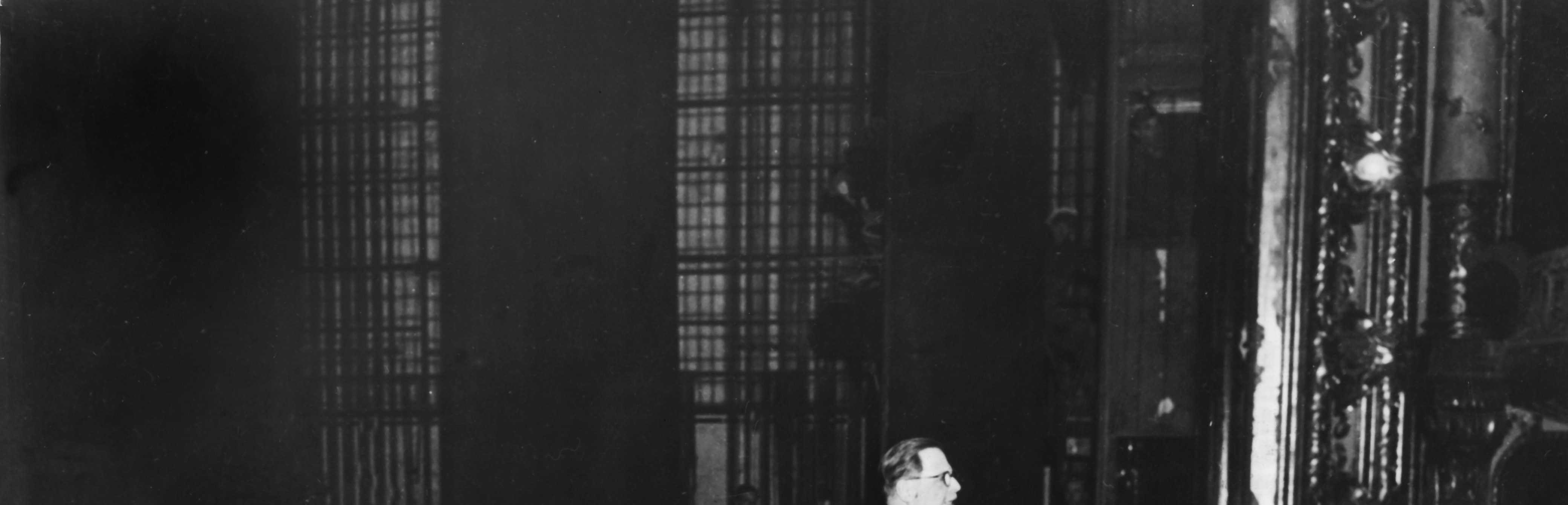 Schwarz-Weiß-Fotografie des Ministerpräsidenten Karl Geiler am Rednerpult mit acht weiteren Ministern hinter ihm sitzend im Jahre 1946
